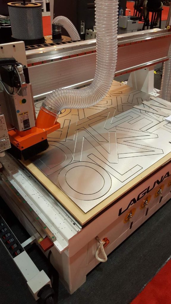 CNC router machine cutting geometric patterns into a sheet with dust collection hose attached.