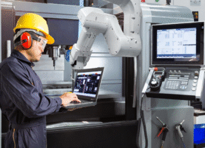 Engineer monitoring CNC machine with robotic arm integration using laptop for industrial automation control
